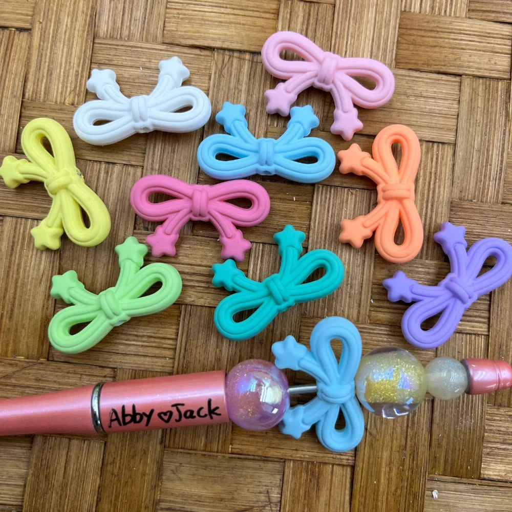 Colorful pastel bow-shaped acrylic beads with star accents in pink, blue, green, purple, orange, teal, yellow, white and more, displayed on a wicker surface next to a pink beadable pen branded Abby Jack.