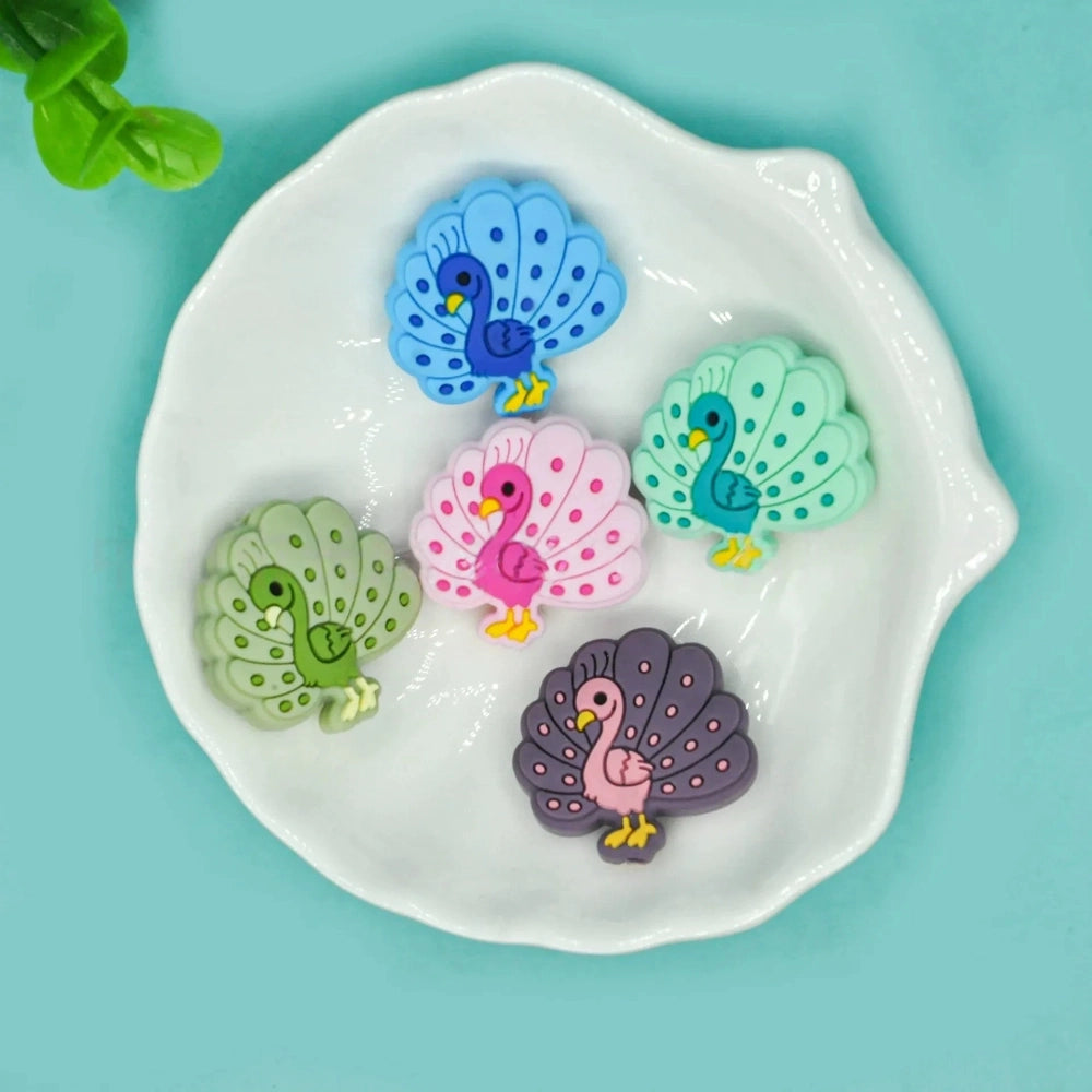 Five colorful peacock silicone focal beads in blue, mint green, olive green, pink, and purple displayed on a white ceramic dish against a teal background.