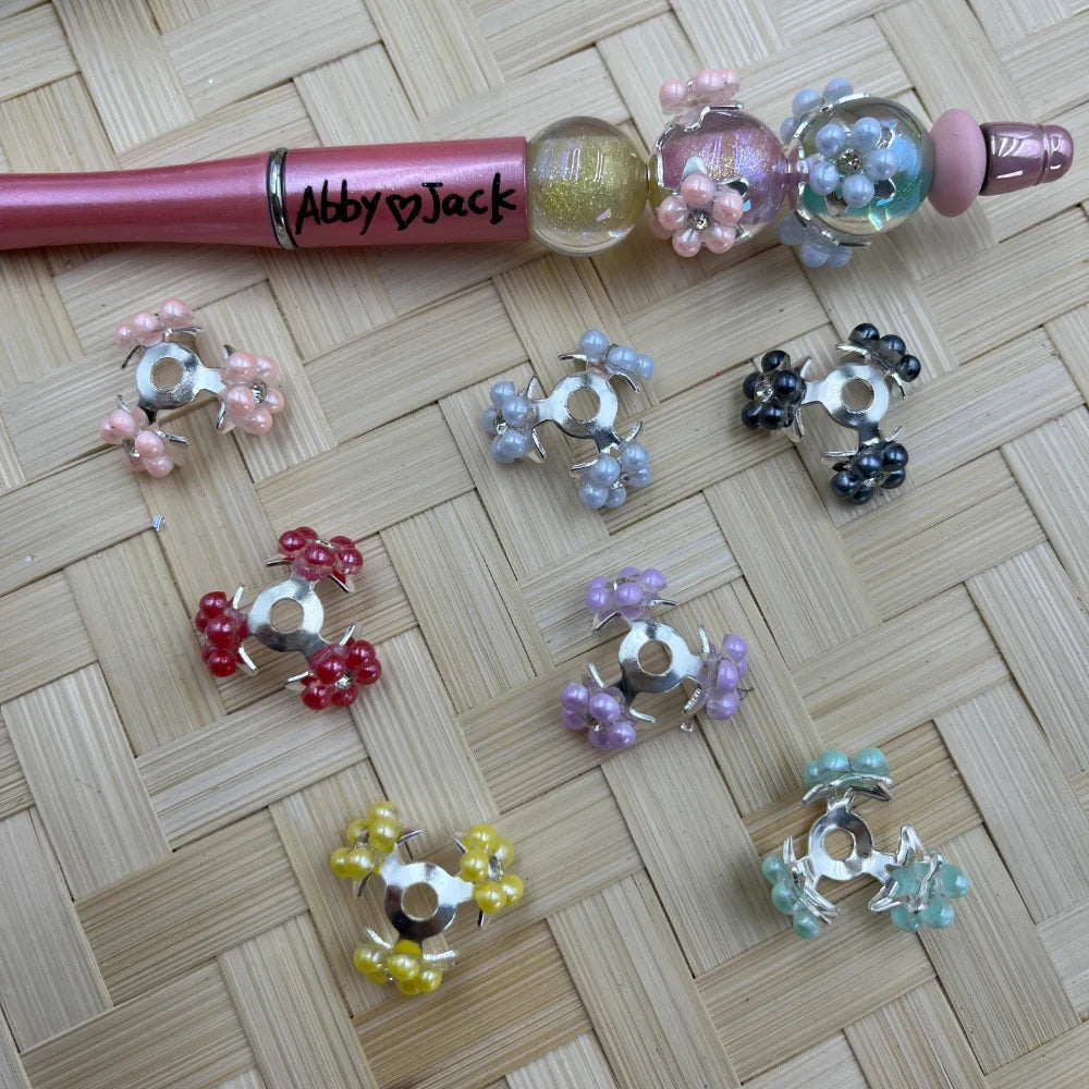 Assorted pearl flower cluster silver bail beads in pink, red, yellow, lavender, mint, gray, and black, displayed on a woven surface next to a decorated beadable pen labeled Abby Jack.