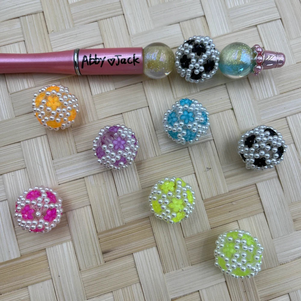 Assorted pearl-rimmed daisy flower bubblegum beads in orange, purple, pink, teal, neon yellow, and black with white pearl borders, displayed on woven bamboo surface next to a pink beadable pen labeled Abby and Jack.