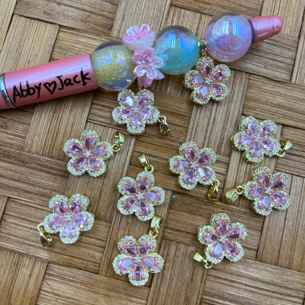 Pink cubic zirconia crystal flower shaped gold bail pendants with pave halo detail, displayed on woven bamboo surface alongside glitter beadable pens.