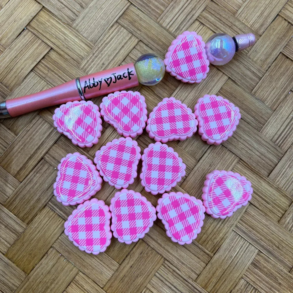 Pink and white gingham plaid Easter egg shaped acrylic beads with scalloped border edges displayed on woven surface next to beadable pens
