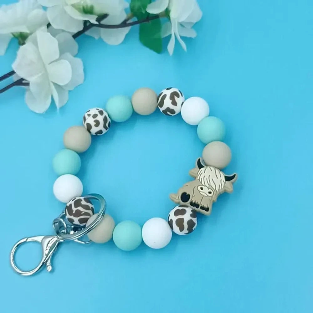 Pink, white, and beige highland cow print silicone beads arranged on a beaded bracelet keychain with a tassel and lobster clasp, displayed on a white and pink surface with green leaves in the background. - Blue