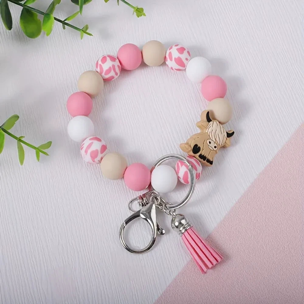 Pink, white, and beige highland cow print silicone beads arranged on a beaded bracelet keychain with a tassel and lobster clasp, displayed on a white and pink surface with green leaves in the background. - Green