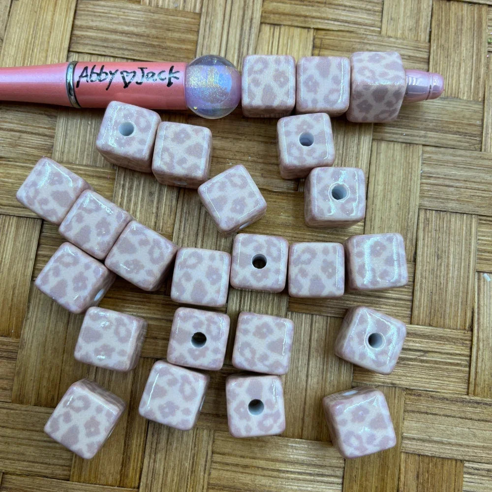 Pink leopard print cube acrylic beads with glossy finish displayed on a woven surface alongside a beadable pen, showing hole detail for DIY jewelry making and beadable pen projects.