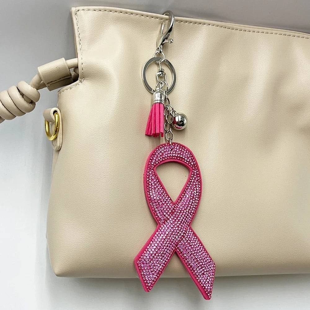 Pink rhinestone-encrusted breast cancer awareness ribbon keychain charm with pink tassel and silver hardware attached to a cream leather handbag.