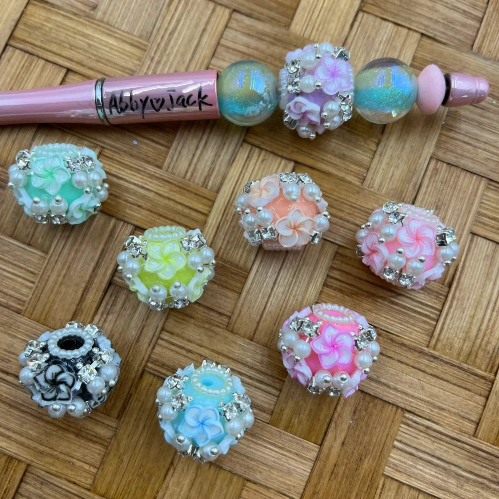 Assorted pastel plumeria flower fancy beads decorated with white pearls and rhinestones in mint, yellow, peach, pink, blue, and black colorways, displayed on woven rattan surface alongside a decorated beadable pen labeled Abby and Jack.