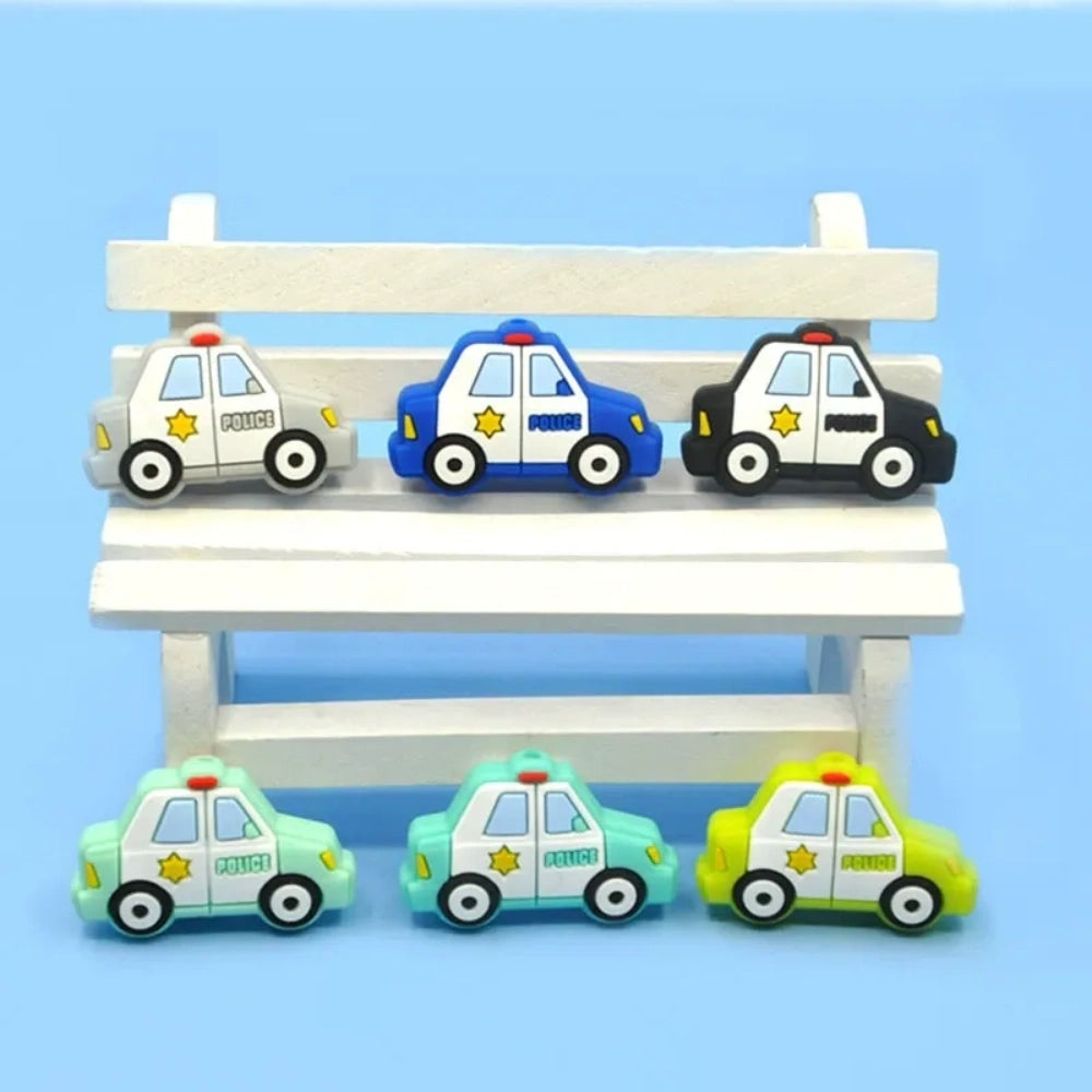 Six cartoon police car silicone focal beads in white, blue, black, mint green, light teal, and yellow displayed on a small wooden bench against a blue background, each featuring a gold sheriff star badge and POLICE text.