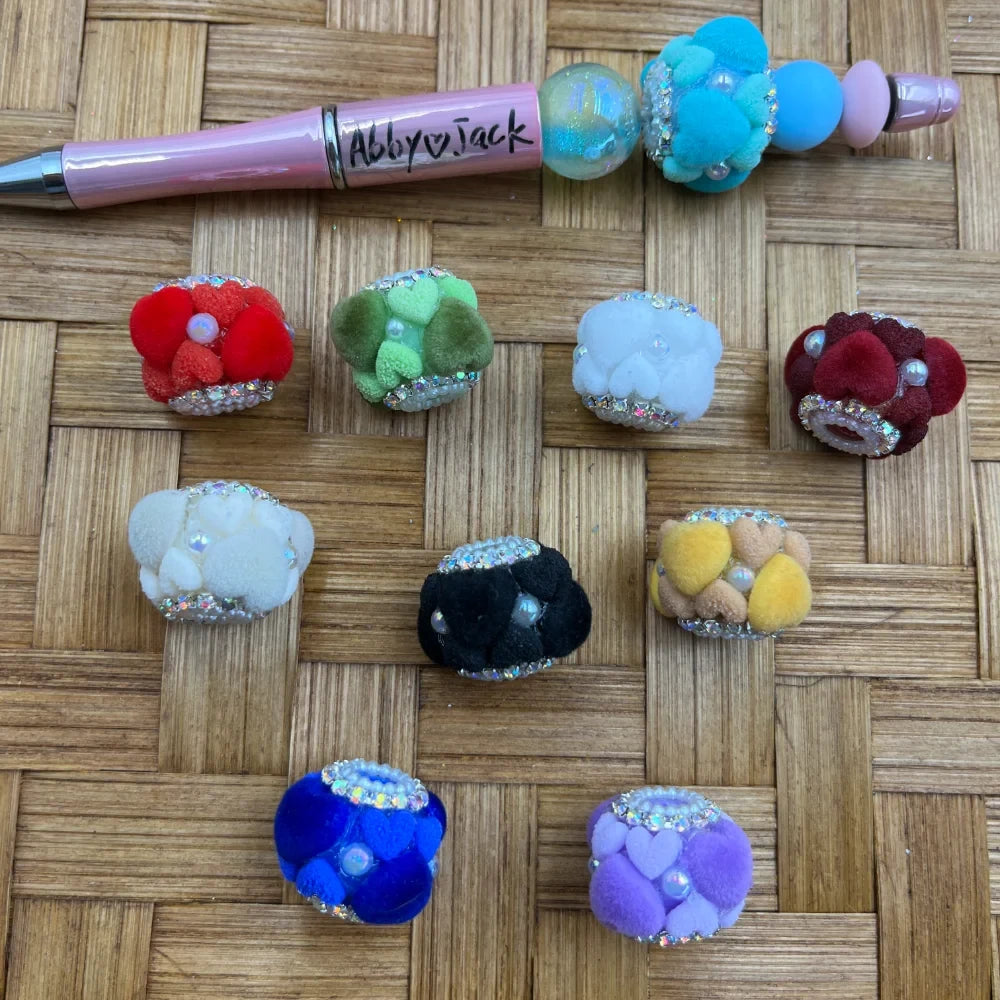 Assorted colorful fluffy pom pom rhinestone fancy beads in red, green, white, dark red, black, yellow, blue, and purple displayed on woven surface with a decorated beadable pen.