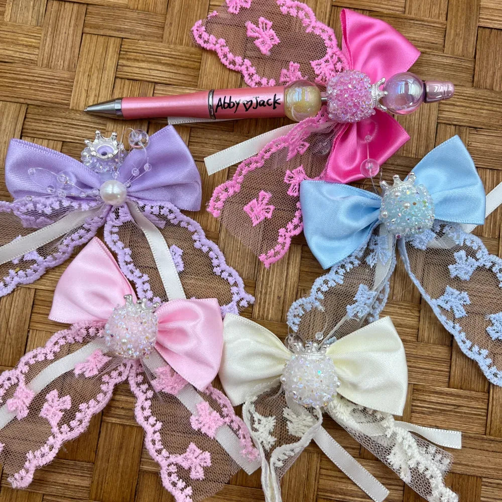 Assorted pastel princess crown bow fancy beads in purple, pink, blue, cream and hot pink with lace trim, pearl and crystal accents displayed on wicker surface alongside a decorated beadable pen.