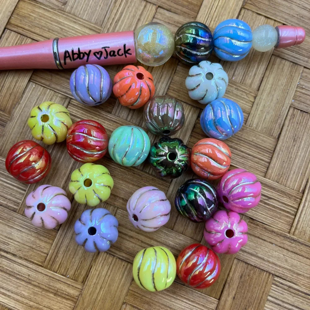 Assorted colorful pumpkin-shaped AB finish acrylic beads in purple, orange, red, yellow, pink, blue, green, and multicolor iridescent finishes displayed on a wicker surface next to a beadable pen labeled Abby and Jack.