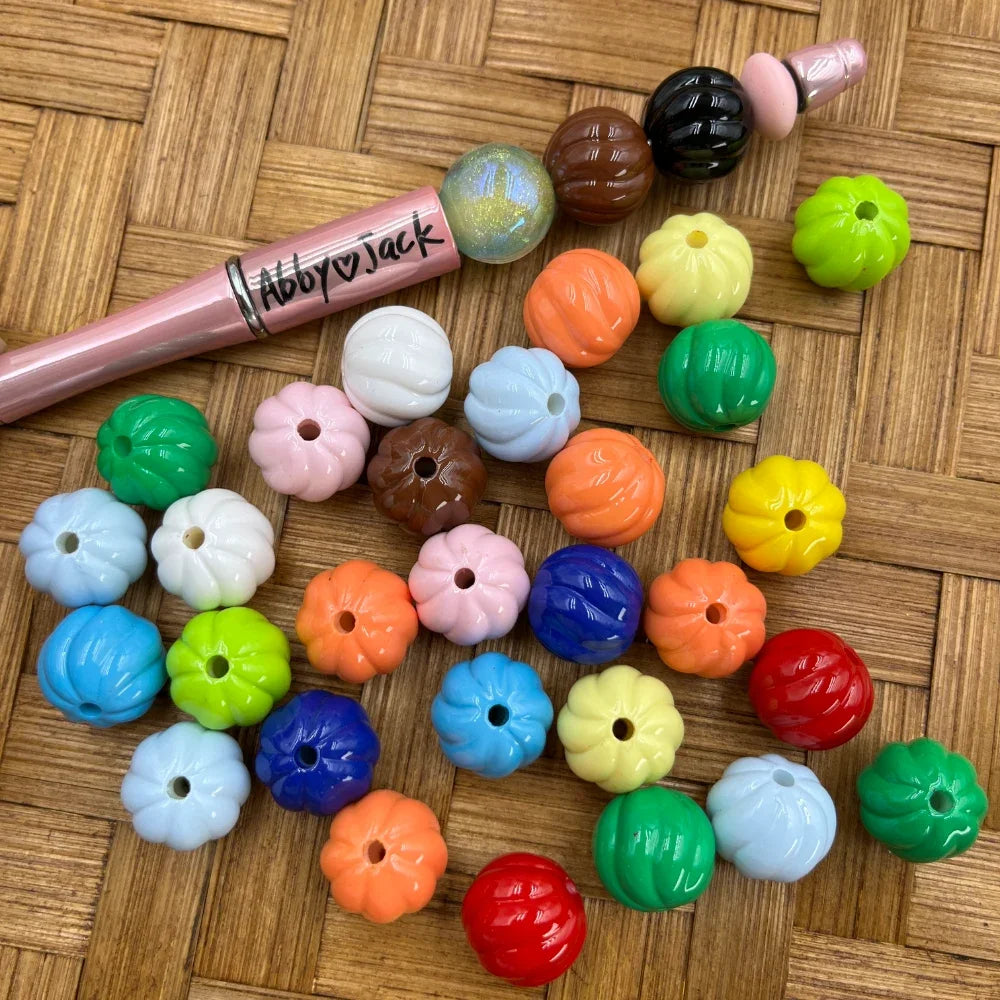 Assorted colorful pumpkin shaped acrylic beads in multiple colors including orange, blue, green, red, yellow, pink, white, and more, displayed on a wicker surface next to a beadable pen.