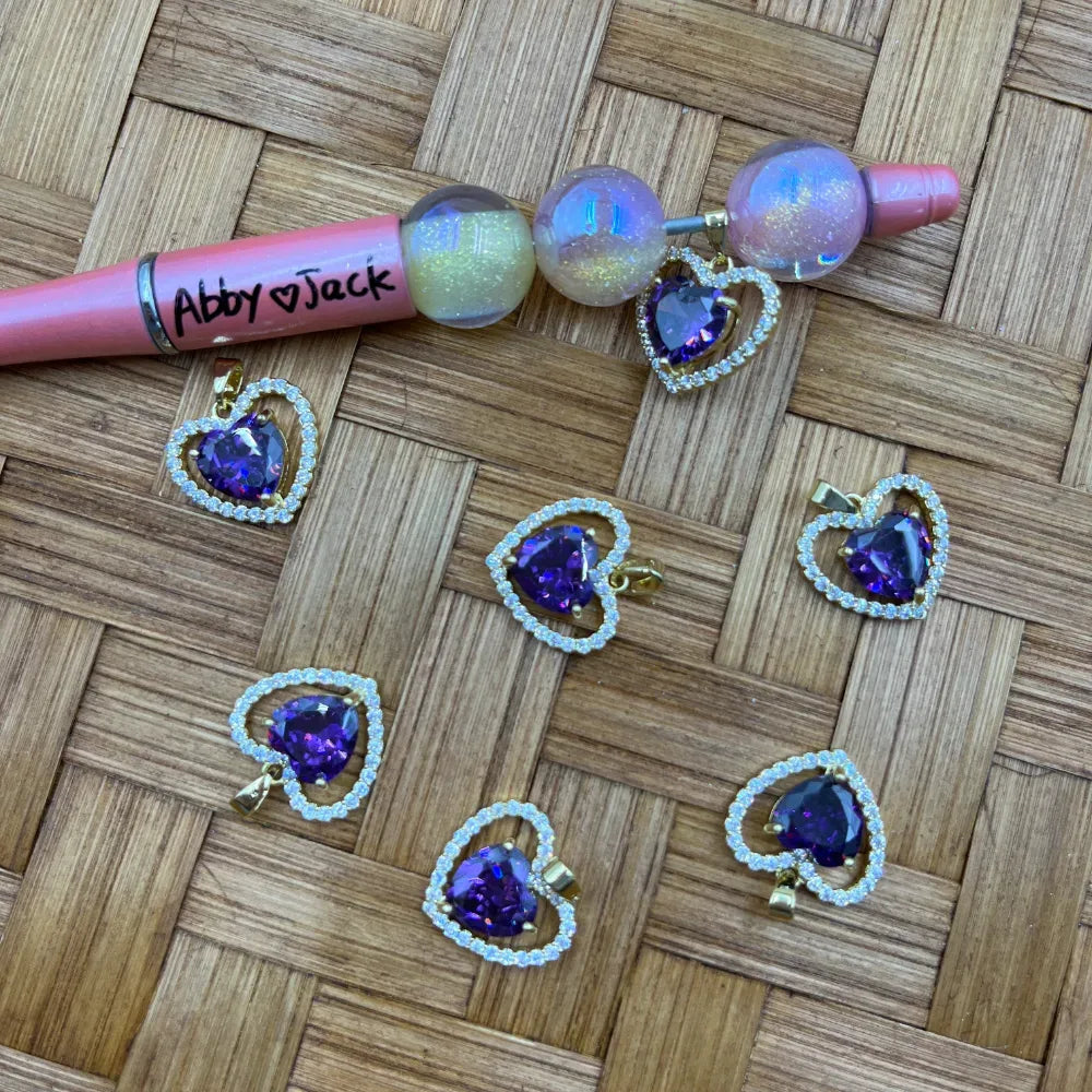 Purple amethyst heart crystal bail beads with gold cubic zirconia halo setting displayed on woven bamboo surface alongside a pink beadable pen decorated with iridescent globe beads and heart charms.