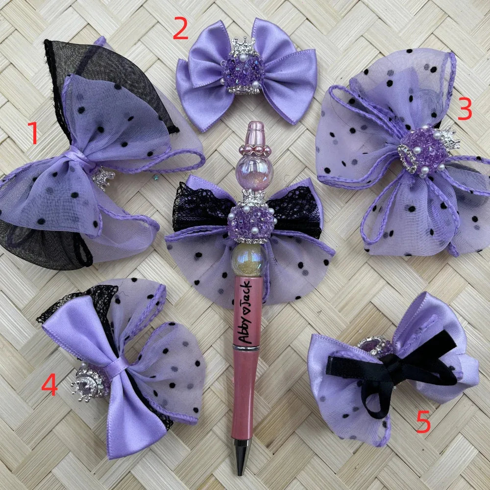 Five purple lavender satin and black polka dot tulle bow pen toppers with rhinestone crown and pearl accents, displayed around a decorated beadable pen labeled Abby Jack on a woven bamboo background.