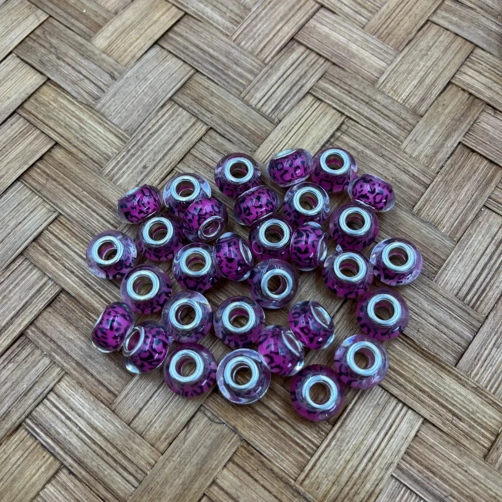 Purple and black leopard print acrylic spacer beads with silver metal core holes, scattered on woven bamboo surface, for beadable pens and DIY jewelry making.