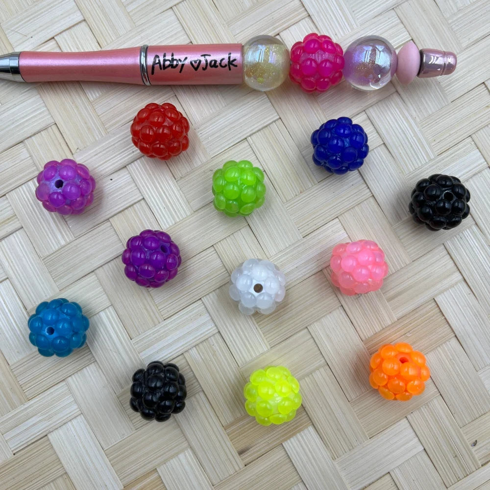 Assorted colorful raspberry berry bumpy acrylic fancy beads in red, purple, blue, green, black, white, pink, teal, orange, and yellow displayed on a woven surface next to a beadable pen.