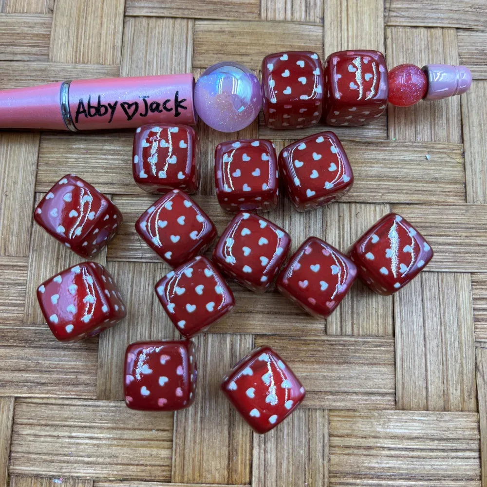 Red cube acrylic beads with white heart pattern scattered on wicker surface next to a beadable pen, perfect for DIY jewelry making and beadable pens.
