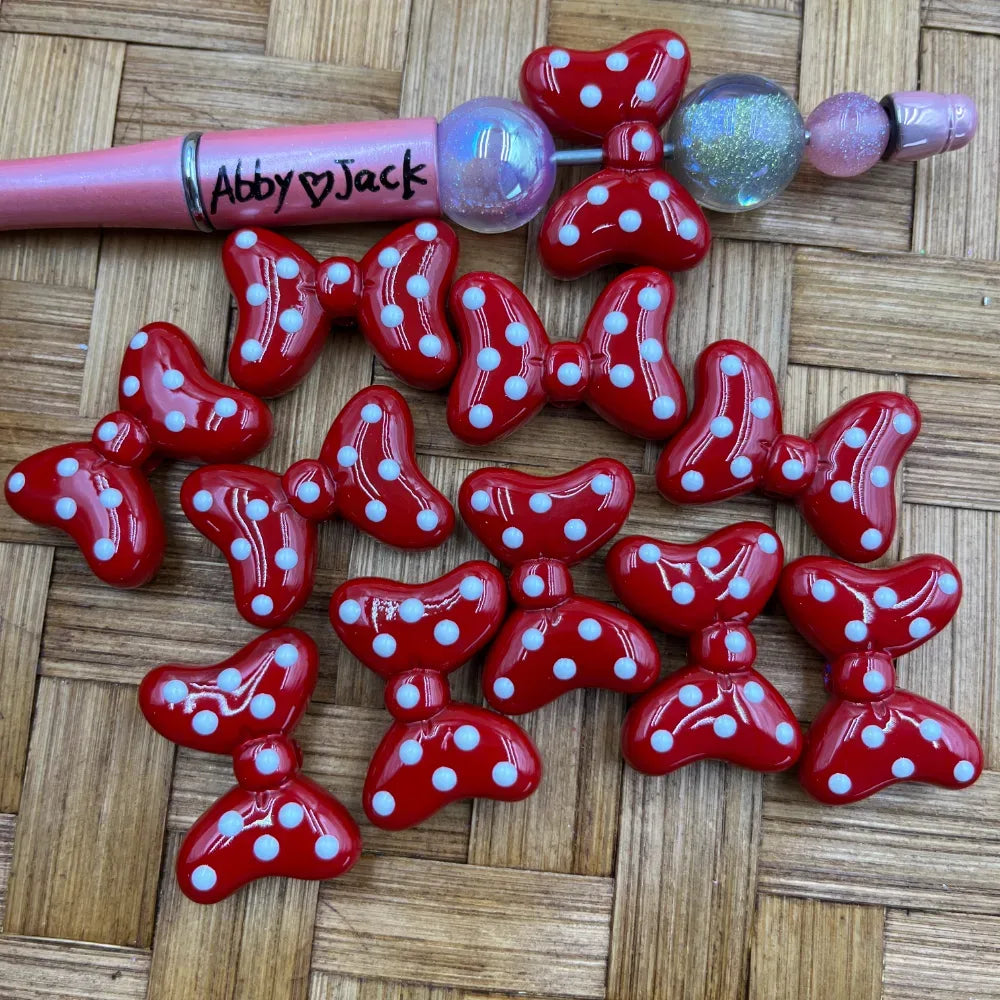 Glossy red and white polka dot bow-shaped acrylic beads displayed on a woven surface next to a pink beadable pen labeled Abby Jack, showing the beads threaded on the pen.
