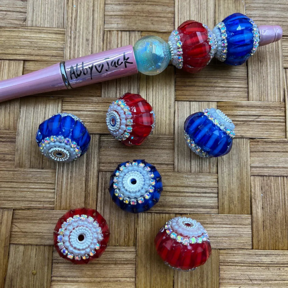 Patriotic red, white, and blue faceted crystal fancy beads with iridescent AB rhinestone trim rings, shown loose and threaded on a pink beadable pen on a wicker surface.