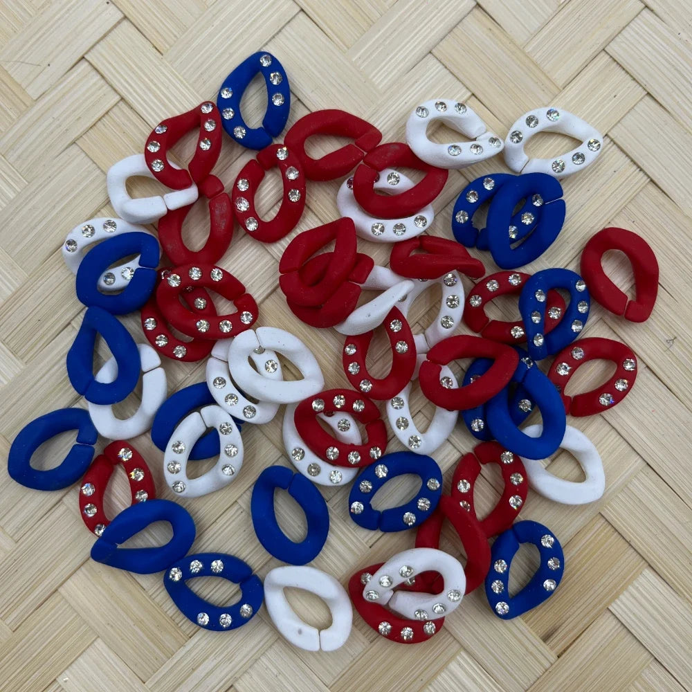 Red, white, and blue oval chain link clay fancy beads embellished with clear rhinestone crystals, scattered on a woven bamboo surface, ideal for patriotic DIY jewelry making and beadable pens.