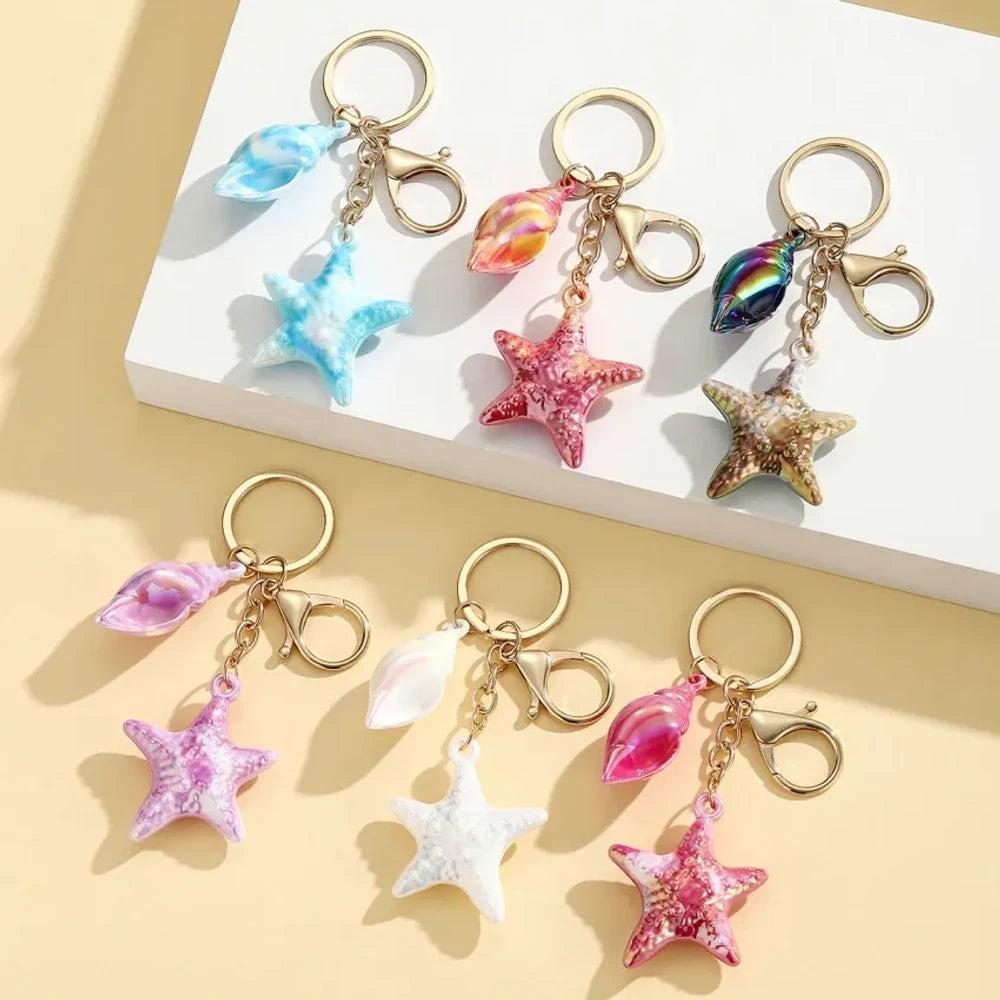 Six colorful resin ocean-themed keychains featuring starfish and seashell charms in blue, pink, purple, white, and iridescent finishes with gold metal rings and lobster clasps.