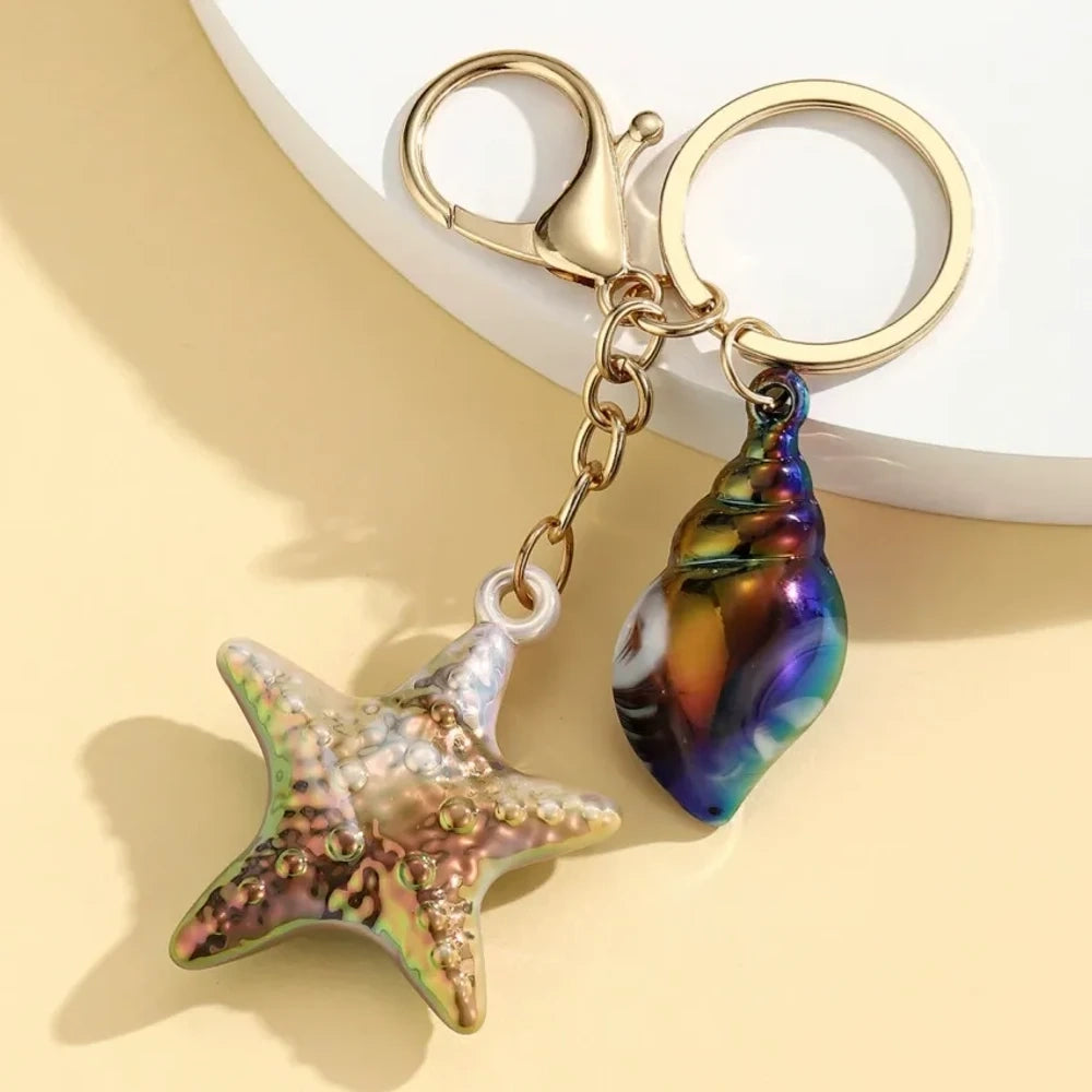Six colorful resin ocean-themed keychains featuring starfish and seashell charms in blue, pink, purple, white, and iridescent finishes with gold metal rings and lobster clasps. - 1