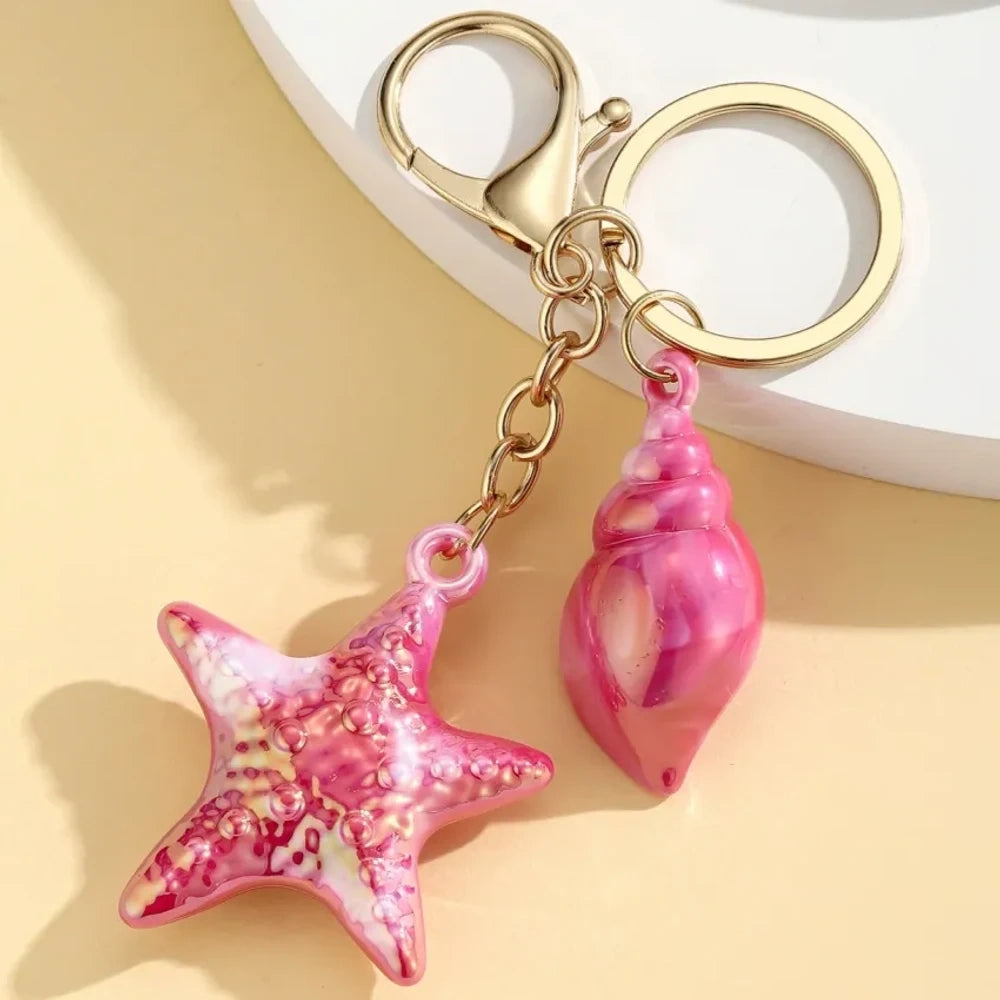 Six colorful resin ocean-themed keychains featuring starfish and seashell charms in blue, pink, purple, white, and iridescent finishes with gold metal rings and lobster clasps. - 2