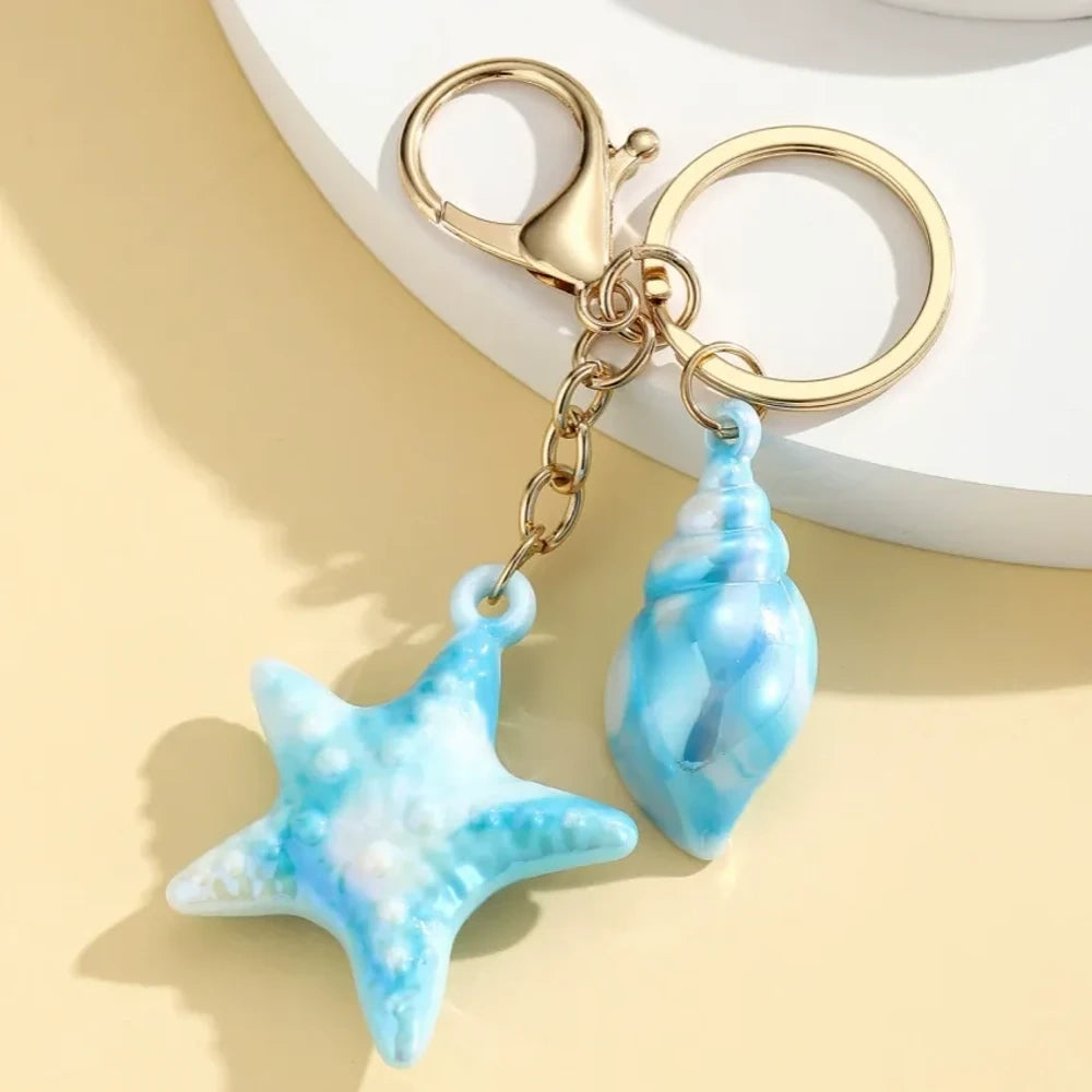 Six colorful resin ocean-themed keychains featuring starfish and seashell charms in blue, pink, purple, white, and iridescent finishes with gold metal rings and lobster clasps. - 3