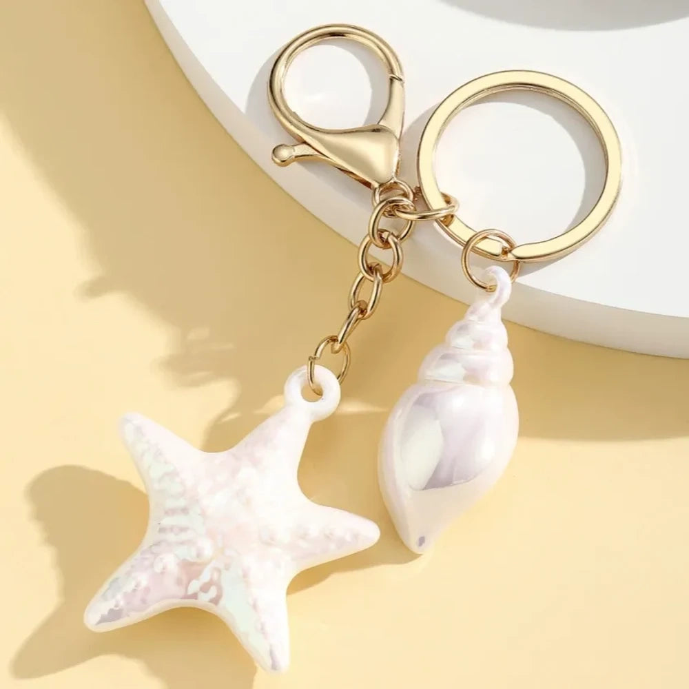 Six colorful resin ocean-themed keychains featuring starfish and seashell charms in blue, pink, purple, white, and iridescent finishes with gold metal rings and lobster clasps. - 4