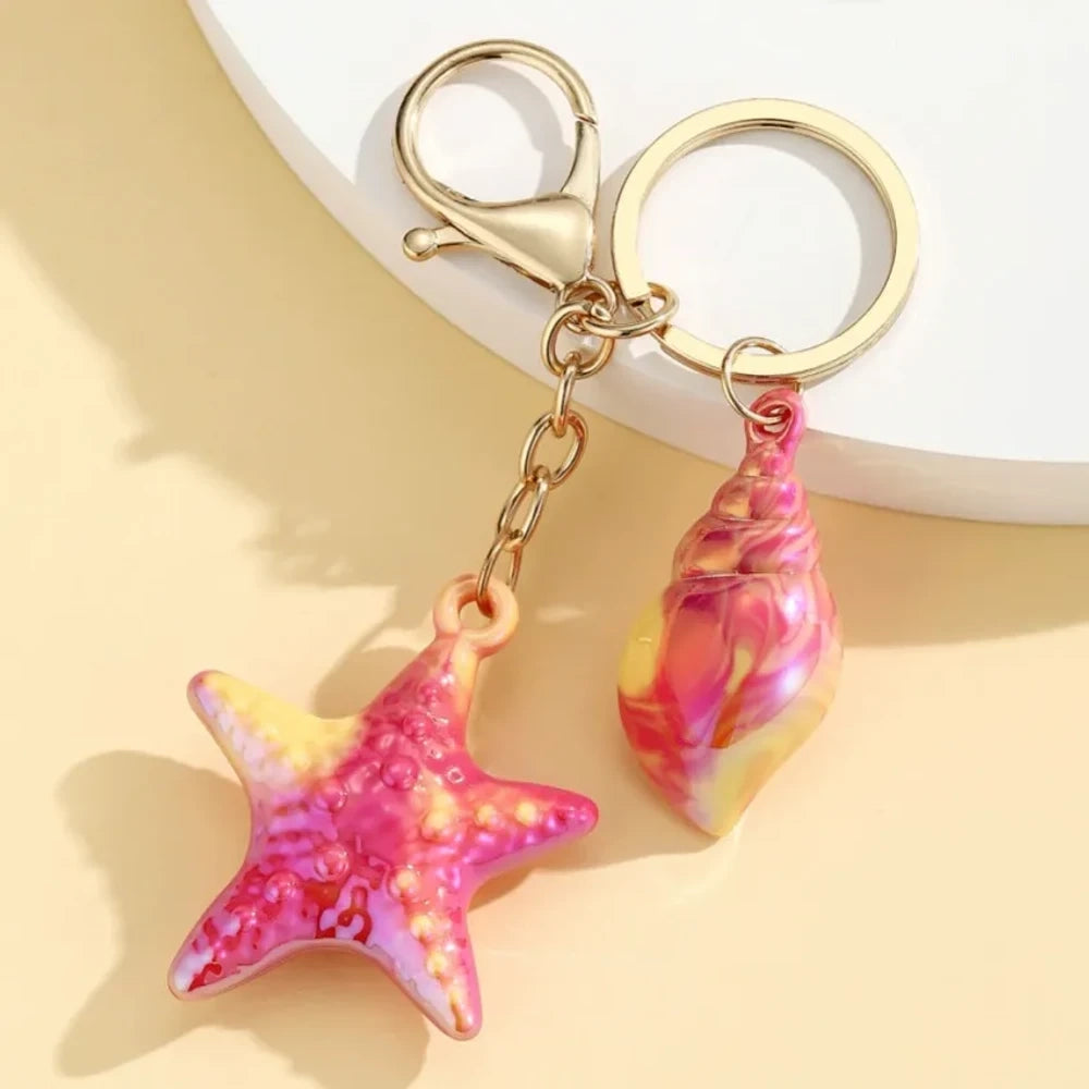 Six colorful resin ocean-themed keychains featuring starfish and seashell charms in blue, pink, purple, white, and iridescent finishes with gold metal rings and lobster clasps. - 5