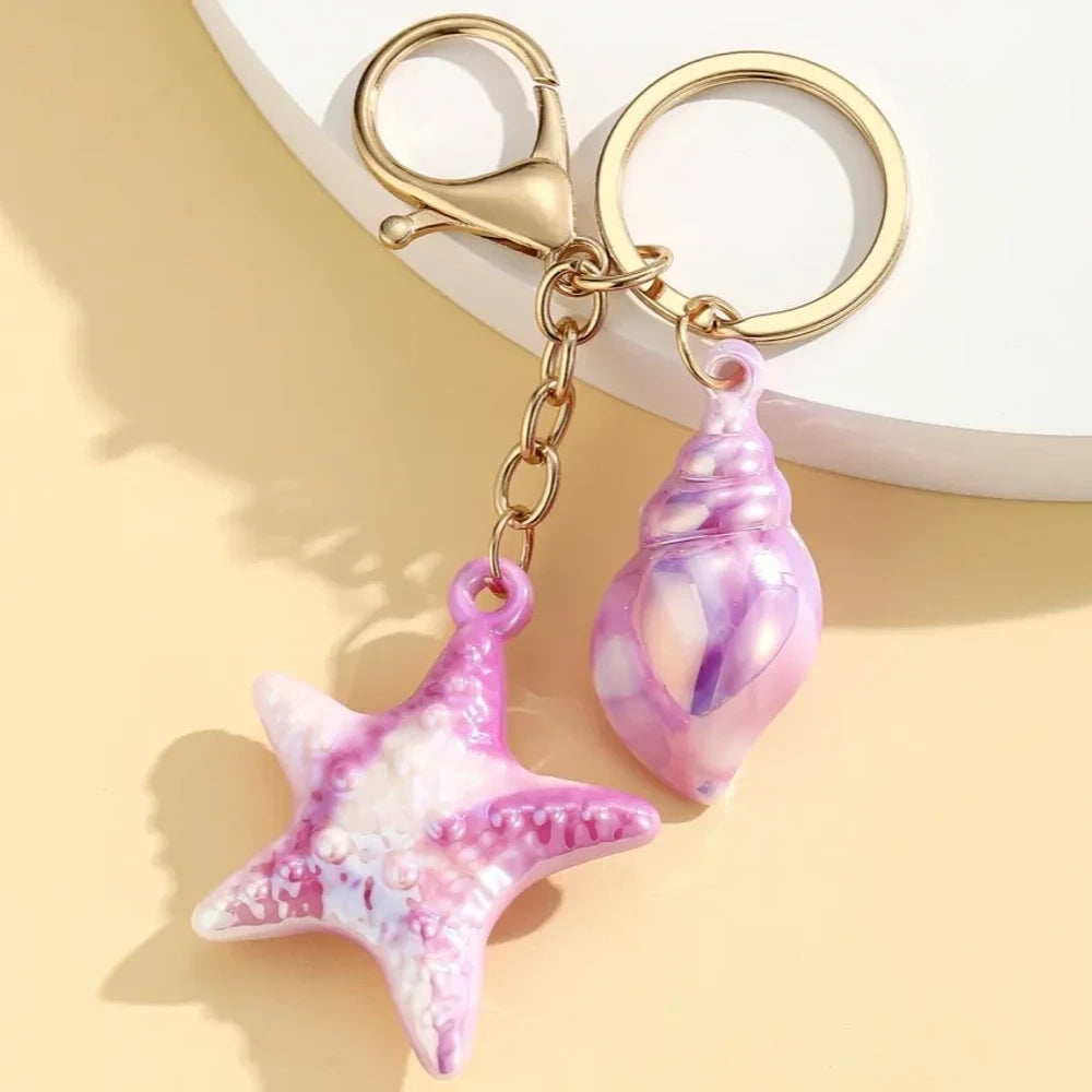 Six colorful resin ocean-themed keychains featuring starfish and seashell charms in blue, pink, purple, white, and iridescent finishes with gold metal rings and lobster clasps. - 6