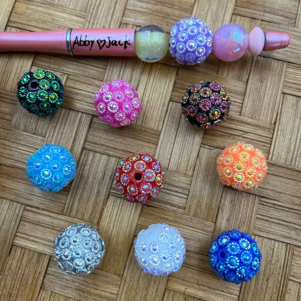 Assorted color rhinestone bling ball beads in teal, pink, black, blue, red, orange, silver, white, and cobalt blue displayed on woven surface next to a beadable pen labeled Abby Jack.