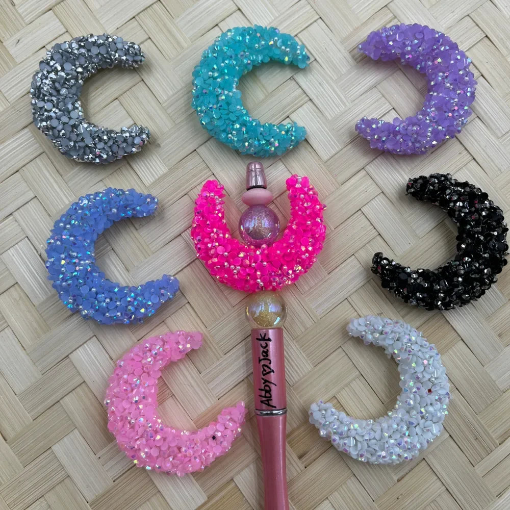 Seven rhinestone-covered crescent moon flatback embellishments in silver, teal, purple, blue, pink, black, and white arranged around a hot pink beadable pen on a wicker background.