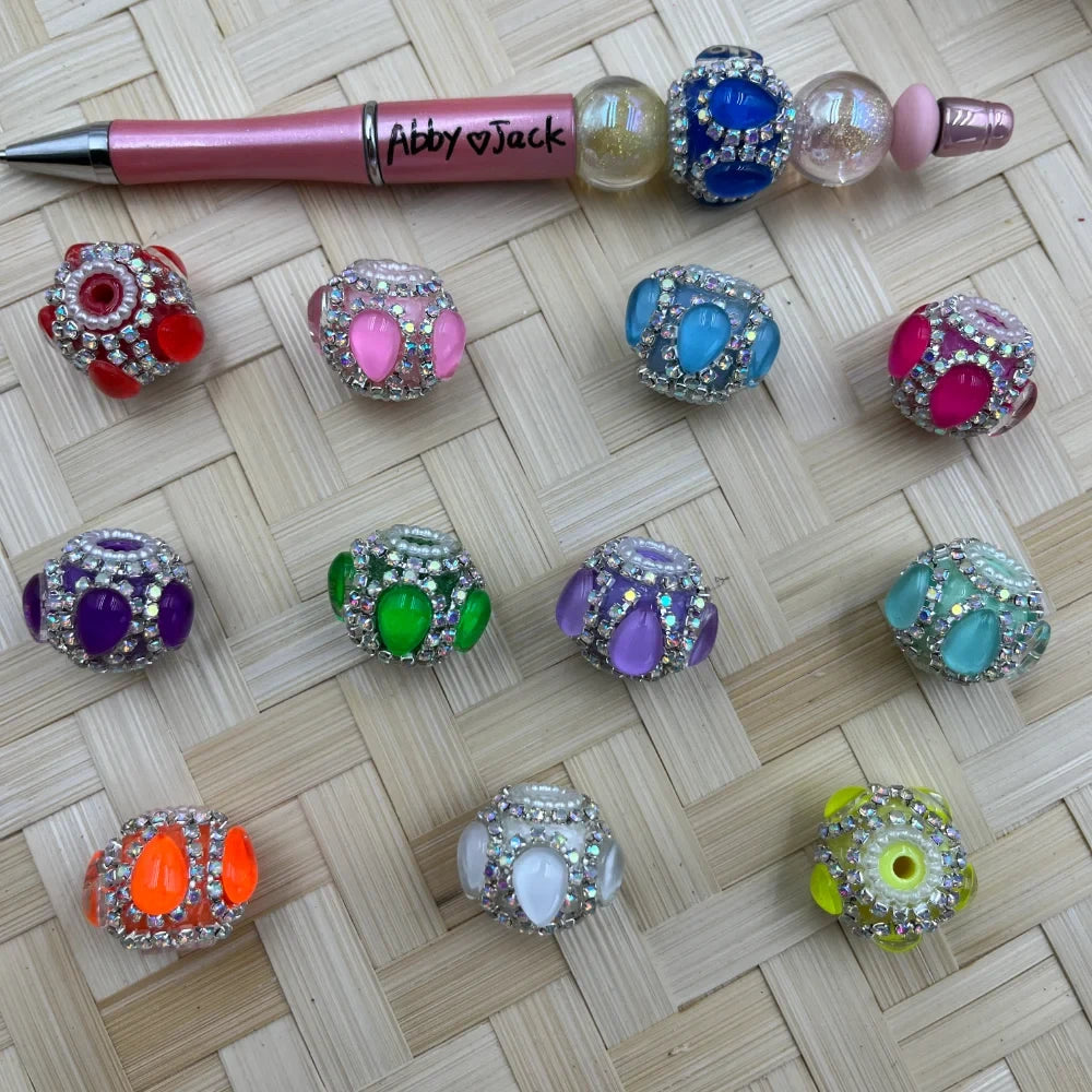 Assorted rhinestone crystal fancy chunky beads in 11 colors including red, pink, blue, purple, green, lavender, teal, orange, white, and yellow-green, displayed alongside a pink beadable pen labeled Abby Jack on a woven bamboo background.