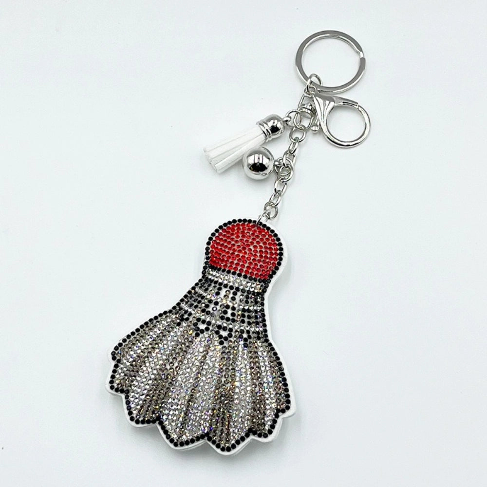 Rhinestone-encrusted badminton shuttlecock keychain charm with red crystal top, silver and black rhinestone feathers, white tassel, and silver key ring on white background.