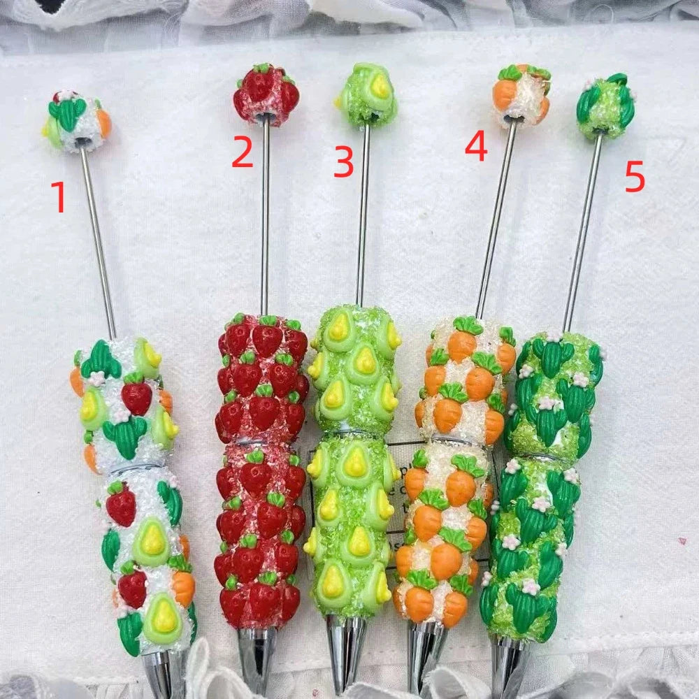 Five rhinestone beadable pens decorated with fruit and vegetable charms including strawberries, avocados, carrots, and cacti on crystal-encrusted handles with silver metal tips.