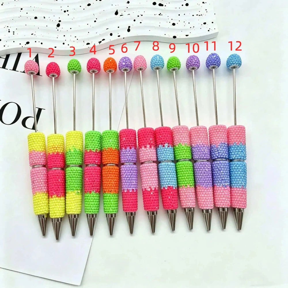 Set of 12 rhinestone beadable pens in multicolor sparkle combinations including pink, yellow, green, orange, purple, and blue beads with silver metal tips, numbered 1 through 12, displayed on a white surface.