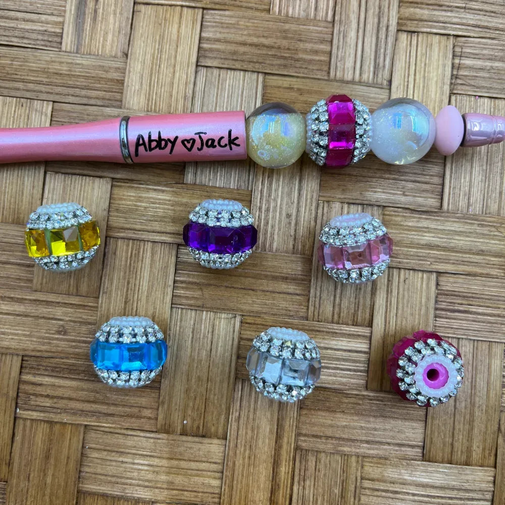 Assorted rhinestone bling acrylic fancy beads in yellow, purple, pink, blue, clear, hot pink, and iridescent colors displayed on a woven surface next to a personalized pink beadable pen labeled Abby and Jack.