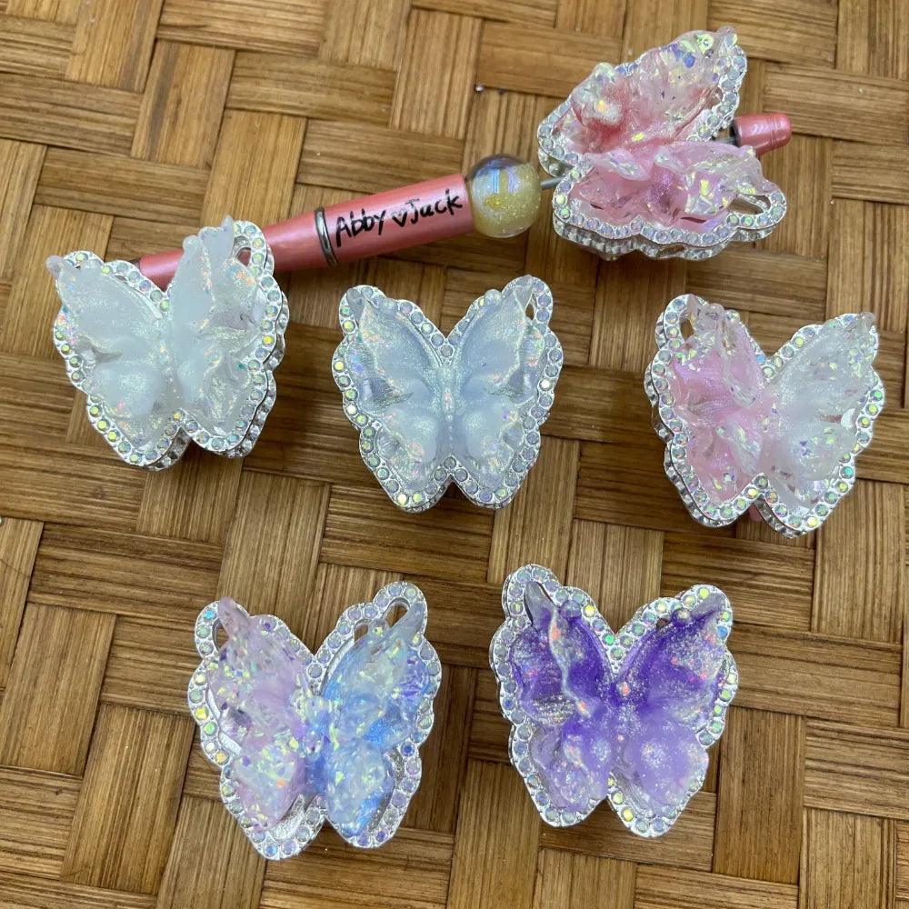 Assorted glitter resin butterfly beads with AB rhinestone borders in white, pink, lavender, and purple, displayed with a beadable pen on a wicker surface.