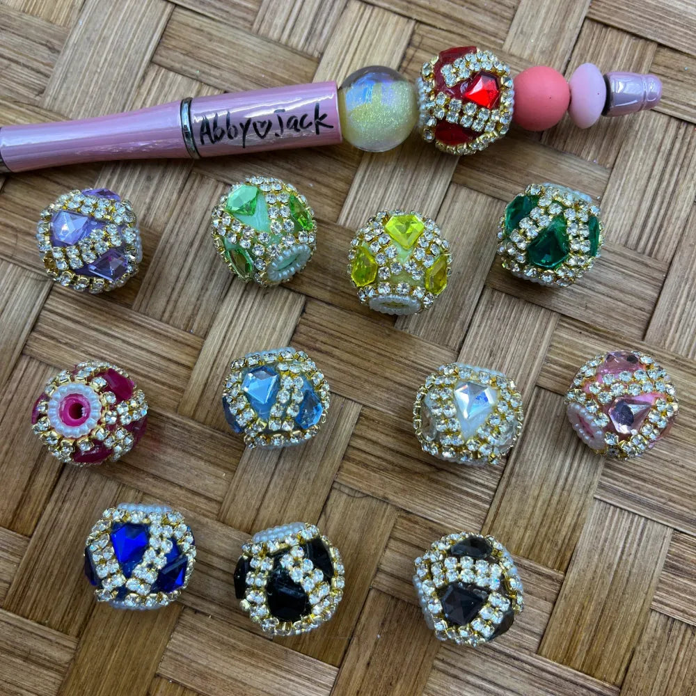 Assorted rhinestone-bordered gemstone fancy beads in purple, green, yellow, teal, red, blue, black, and pink displayed on a woven surface alongside a decorated beadable pen labeled Abby and Jack.