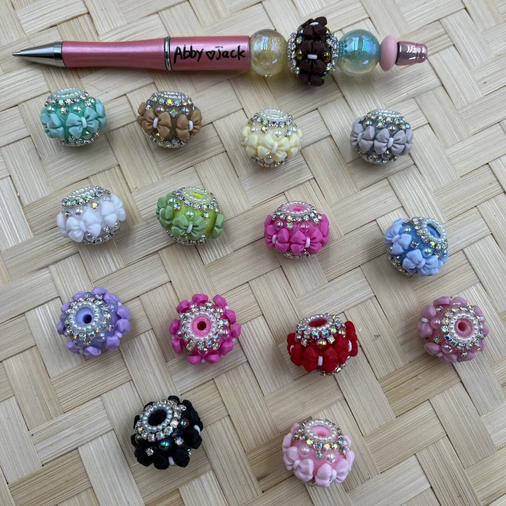 Assorted rhinestone-trimmed clay bow and flower fancy beads in multiple colors including pink, green, purple, red, black, and blue, displayed on a woven surface alongside a beadable pen.