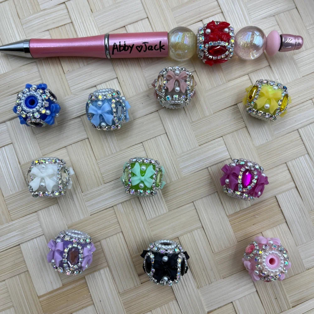 Assorted rhinestone-encrusted fancy beads with bow and flower embellishments in multiple colors including blue, pink, green, black, purple, yellow, and white, displayed next to a decorated beadable pen on a woven surface.