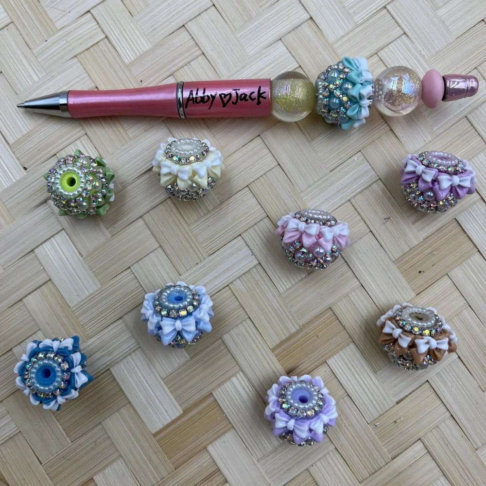 Assorted fancy beads decorated with rhinestones, bows, and pearls in pastel colors including blue, pink, purple, and brown, displayed next to a pink beadable pen on a woven bamboo surface.