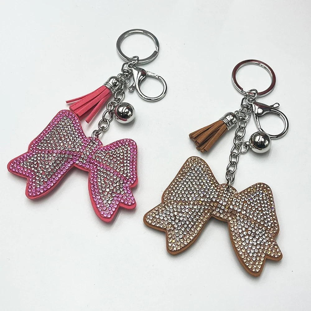 Two rhinestone-encrusted bow keychains with leather tassels and silver bell pendants in pink and brown colorways on a white background.