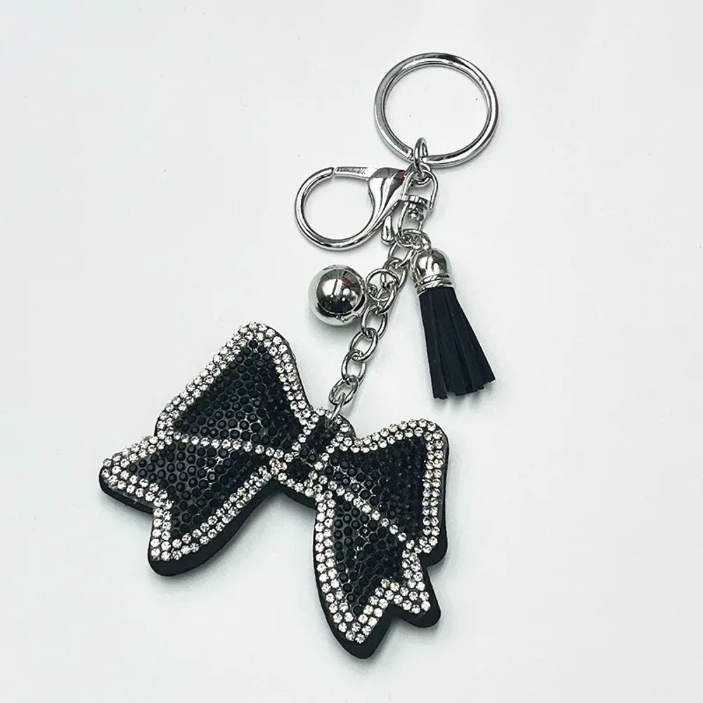 Two rhinestone-encrusted bow keychains with leather tassels and silver bell pendants in pink and brown colorways on a white background. - Black