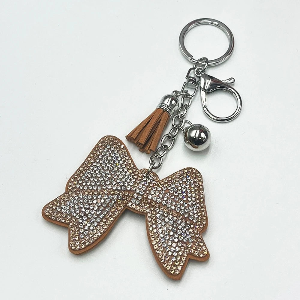 Two rhinestone-encrusted bow keychains with leather tassels and silver bell pendants in pink and brown colorways on a white background. - Khaki