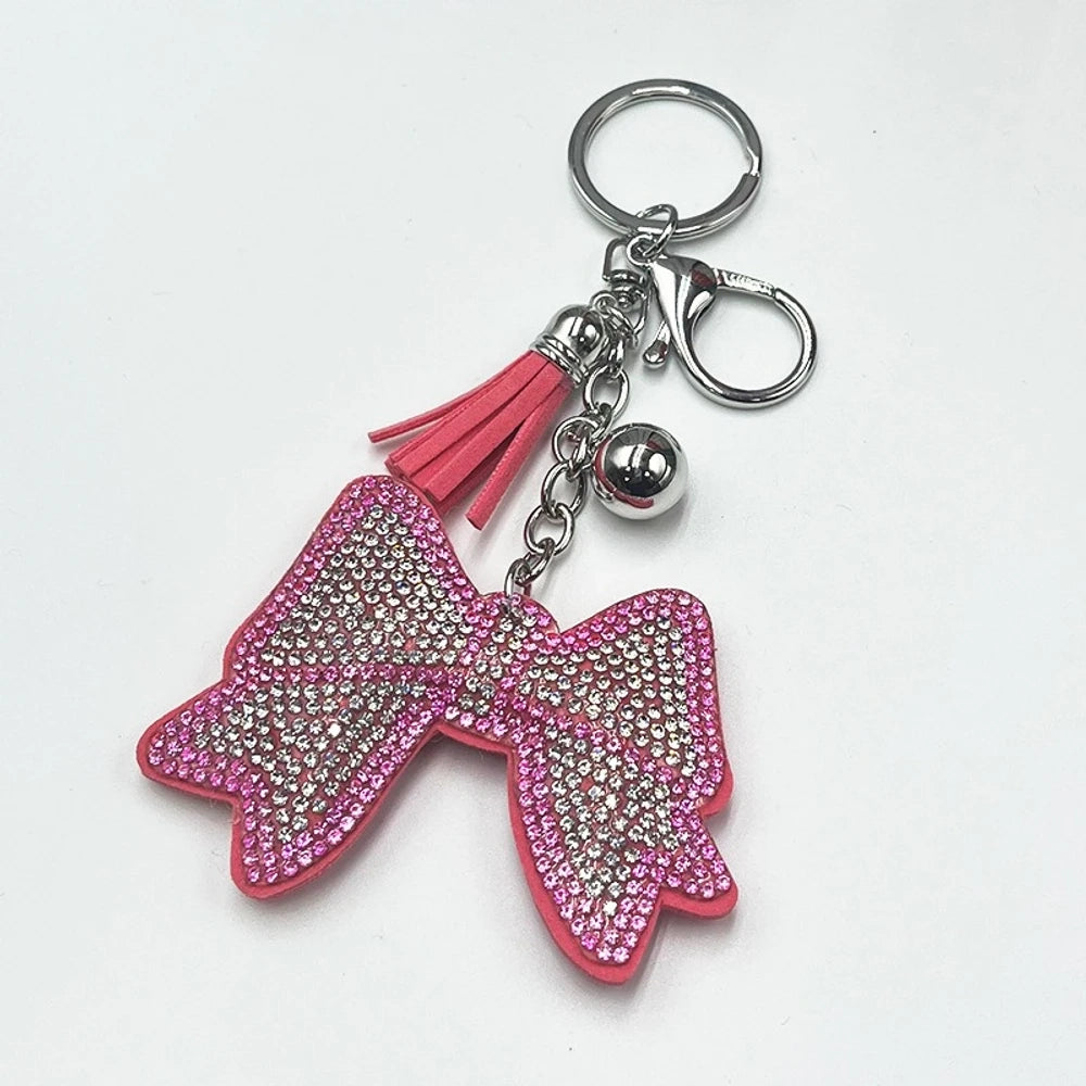 Two rhinestone-encrusted bow keychains with leather tassels and silver bell pendants in pink and brown colorways on a white background. - Pink