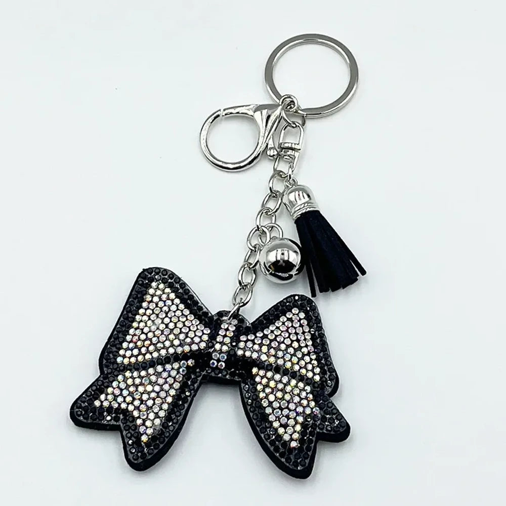 Two rhinestone-encrusted bow keychains with leather tassels and silver bell pendants in pink and brown colorways on a white background. - White