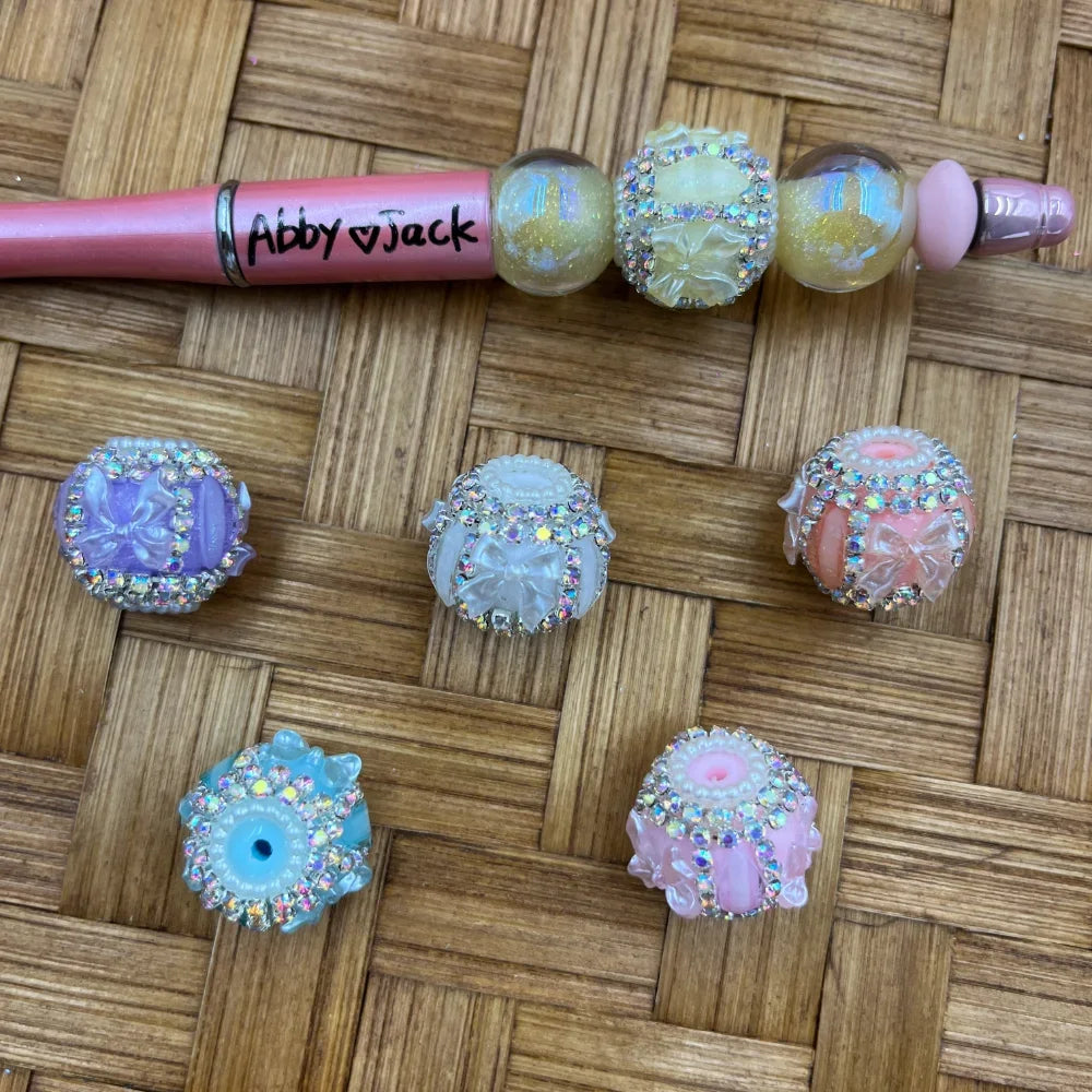 Assorted pastel rhinestone-bordered bow resin fancy beads in purple, white, peach, blue, and pink, displayed on a woven surface next to a pink beadable pen labeled Abby Jack.