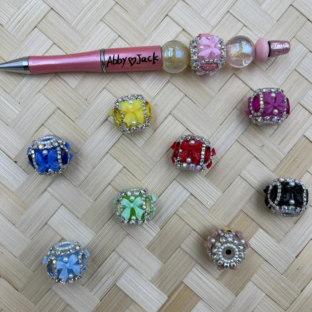 Assorted colorful square rhinestone-bordered beads featuring 3D bow centerpieces in yellow, blue, red, green, pink, magenta, black, and light blue, displayed on wicker surface alongside a decorated beadable pen labeled Abby Jack.
